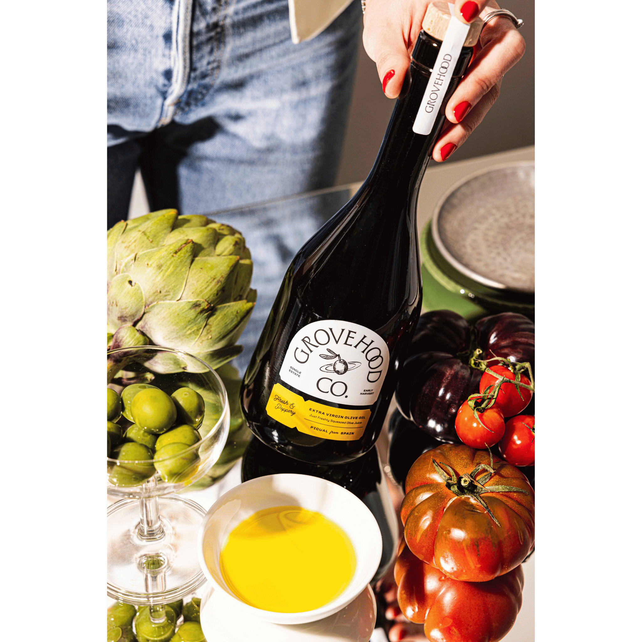 Grovehood Collective Spanish extra virgin olive oil bottle at a table with tomatoes, olives and artichoke